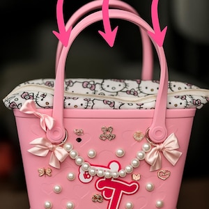 Peut inclure: Sac fourre-tout rose avec une doublure imprimée Hello Kitty et des embellissements décoratifs. Comprend des perles, des nœuds et des breloques dorées. Le sac est de forme rectangulaire avec une poignée et le mot "bogg" en bas.