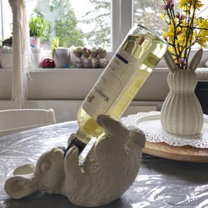 May include: White ceramic bunny bottle holder with a bottle of white wine inside. The bottle label reads "Marcoux" and "Vin Blanc".