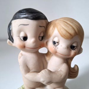 Può includere: Statuina vintage in ceramica raffigurante una coppia che si abbraccia. Le figure hanno guance rosee, capelli neri e sono nude. La donna ha lentiggini e capelli biondi. La statuina è un pezzo romantico e sentimentale.