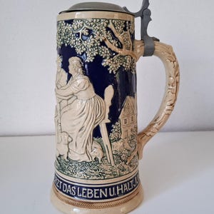 Peut inclure: Une chope à bière en céramique avec un couvercle à charnière en métal et une anse en forme de branche d'arbre. La chope présente un motif en relief d'un couple, d'un chien et d'une maison, avec les mots "Nutz das Leben u. Halt" autour de la base. Les couleurs sont crème, bleu et vert.