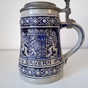 Op de afbeelding: Een blauw-witte keramische bierpul met een metalen scharnierend deksel. De pul heeft een gedetailleerd reliëfontwerp met het woord "BAYERN" en een wapenschild. De handgreep is gebogen en de pul heeft een decoratief patroon.