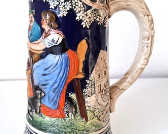 May include: A ceramic beer stein with a detailed relief scene of people, dogs, and a building. The handle is shaped like a tree branch. The stein has a beige base and rim, with a dark blue background.