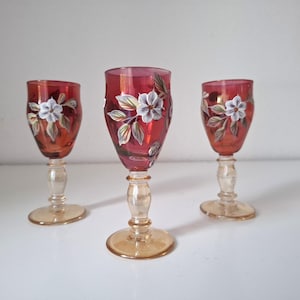 May include: Three small stemmed glasses with deep red bowls featuring hand-painted white flowers and green leaves. Each glass has a clear, amber-tinted stem and a circular base, standing upright in a row against a plain white background.