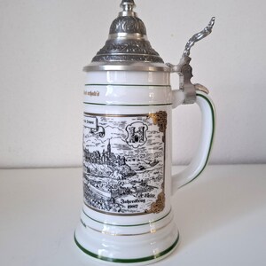 Peut inclure: Chope à bière en céramique blanche avec un couvercle et une poignée en métal argenté. La chope présente une illustration en noir et blanc d'une ville, avec des accents dorés et une bordure verte. Le texte "Jahrestrug 1987" est visible.