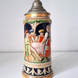 Peut inclure: Une chope à bière en céramique vintage avec une scène en relief détaillée de personnes à une table, avec des personnages en tenue traditionnelle. La chope a un couvercle en métal et une base décorative avec un bord doré. Les couleurs incluent le bleu, le vert, le rouge et le beige.