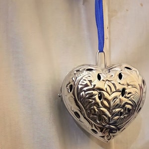 May include: A silver heart-shaped incense burner with intricate floral designs hangs from a blue ribbon. The burner has several small holes for incense sticks.