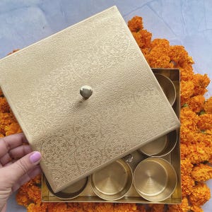 Pode incluir: Uma caixa de metal dourado com uma tampa decorativa e seis compartimentos. A tampa tem um design floral intrincado e uma ma&ccedil;aneta no topo. A caixa &eacute; cercada por flores de cal&ecirc;ndula laranja.