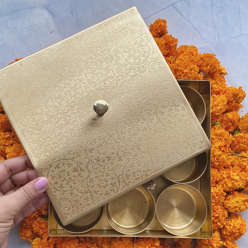 Pode incluir: Uma caixa de metal dourado com uma tampa decorativa e seis compartimentos. A tampa tem um design floral intrincado e uma ma&ccedil;aneta no topo. A caixa &eacute; cercada por flores de cal&ecirc;ndula laranja.