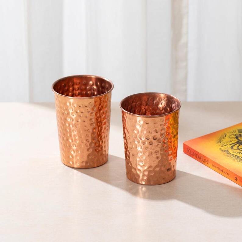 May include: Two copper hammered drinking glasses, one on top of the other, on a white surface.