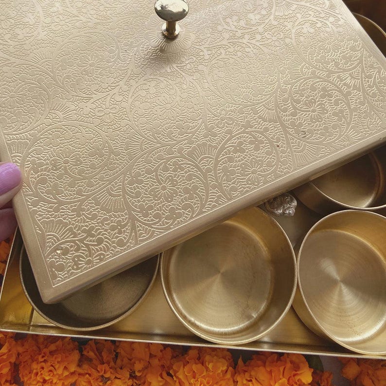 Pode incluir: Uma caixa de metal dourado com uma tampa articulada e padr&otilde;es florais intrincados. A caixa tem seis compartimentos, cada um com um interior liso e arredondado.