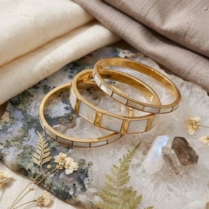 Op de afbeelding: Drie goudkleurige metalen armbanden met rechthoekige inlays van wit parelmoer, gerangschikt in een herhalend patroon rond de omtrek van elke armband. De sieraden zijn cirkelvormig en hebben een gepolijste, reflecterende metalen afwerking.