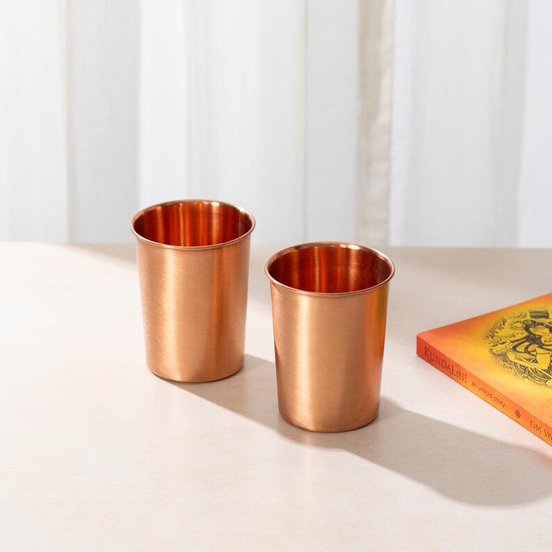 May include: Two copper drinking glasses on a light brown surface. The glasses are made of copper and have a shiny finish.
