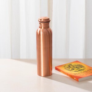 May include: A copper water bottle with a lid. The bottle is on a light brown surface next to a book with the title "Kundalini" on the cover.
