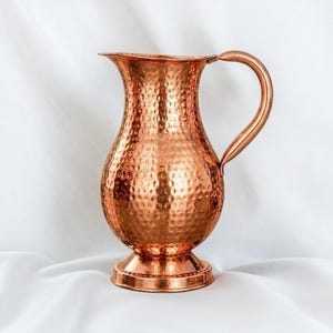 Könnte beinhalten: Ein polierter Kupferkrug mit einer ausgeprägten gehämmerten Textur auf dem abgerundeten Körper. Das Gefäß verfügt über einen breiten, ausgestellten Rand, einen geschwungenen Griff an der Seite und einen runden Sockelfuß, der für Stabilität sorgt.