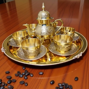 May include: A gold-colored tea set with intricate floral patterns. The set includes a teapot, four cups, saucers, and a serving tray. Coffee beans are scattered on the wooden surface. The tea set is designed with a vintage aesthetic.