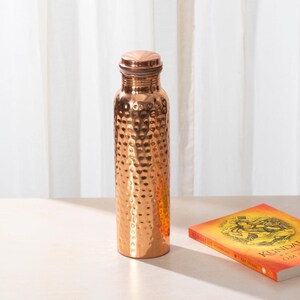 May include: A hammered copper water bottle with a copper lid. The bottle is on a light wood surface.