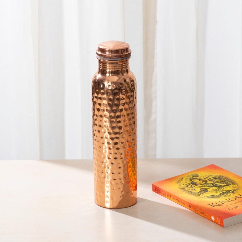 May include: A hammered copper water bottle with a copper lid. The bottle is on a light wood surface.