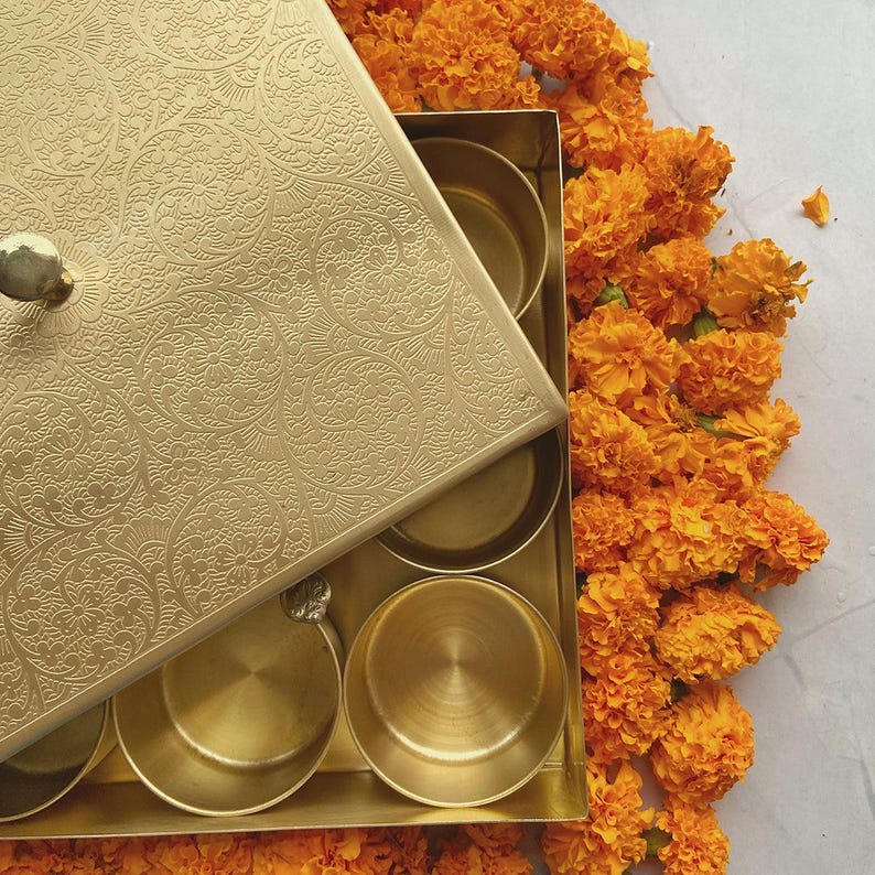 Pode incluir: Uma caixa de metal dourado com um design ornamentado na tampa. A caixa est&aacute; aberta e tem seis compartimentos no interior. A caixa est&aacute; rodeada de flores de cal&ecirc;ndula laranja.