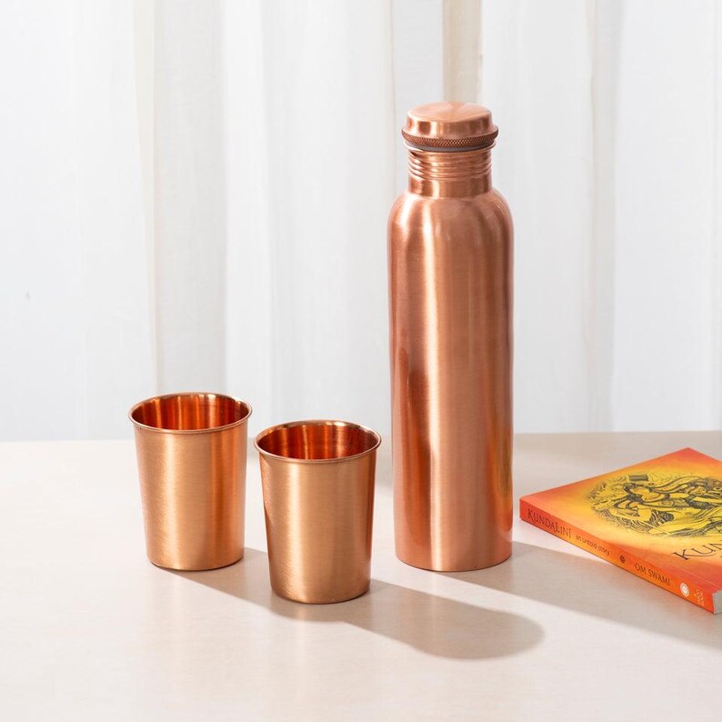May include: Three copper drinking vessels, including a tall bottle and two shorter cups. The bottle has a rounded top and a smooth, shiny surface. The cups are also smooth and shiny. The vessels are arranged on a light-colored surface.