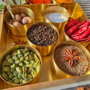 Pode incluir: Uma caixa de especiarias de lat&atilde;o com sete compartimentos, cada um cheio de uma especiaria diferente. As especiarias incluem gr&atilde;os de pimenta preta, vagens de cardamomo, sementes de cominho, anis estrelado, vagens de cardamomo verde, noz-moscada e cravo.