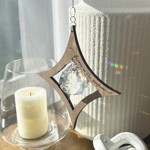 K&ouml;nnte beinhalten: Ein h&ouml;lzerner sternf&ouml;rmiger Kristall-Sonnenf&auml;nger mit dem Text "Forever in the sunshine", der an einer silbernen Kette h&auml;ngt. Der Sonnenf&auml;nger h&auml;ngt vor einer wei&szlig;en, strukturierten Vase und einem wei&szlig;en Keramik-Knoten.