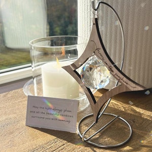 K&ouml;nnte beinhalten: Ein sternf&ouml;rmiger Kristall-Sonnenf&auml;nger aus Holz mit dem eingravierten Text "forever in the sunshine". Der Sonnenf&auml;nger h&auml;ngt an einem Metallst&auml;nder und hat einen Kristall-Prisma, der in der Mitte h&auml;ngt. Eine kleine Karte mit dem Text "May the light always shine and all the beautiful rainbows surround you with memories." liegt auf dem Tisch unter dem Sonnenf&auml;nger.