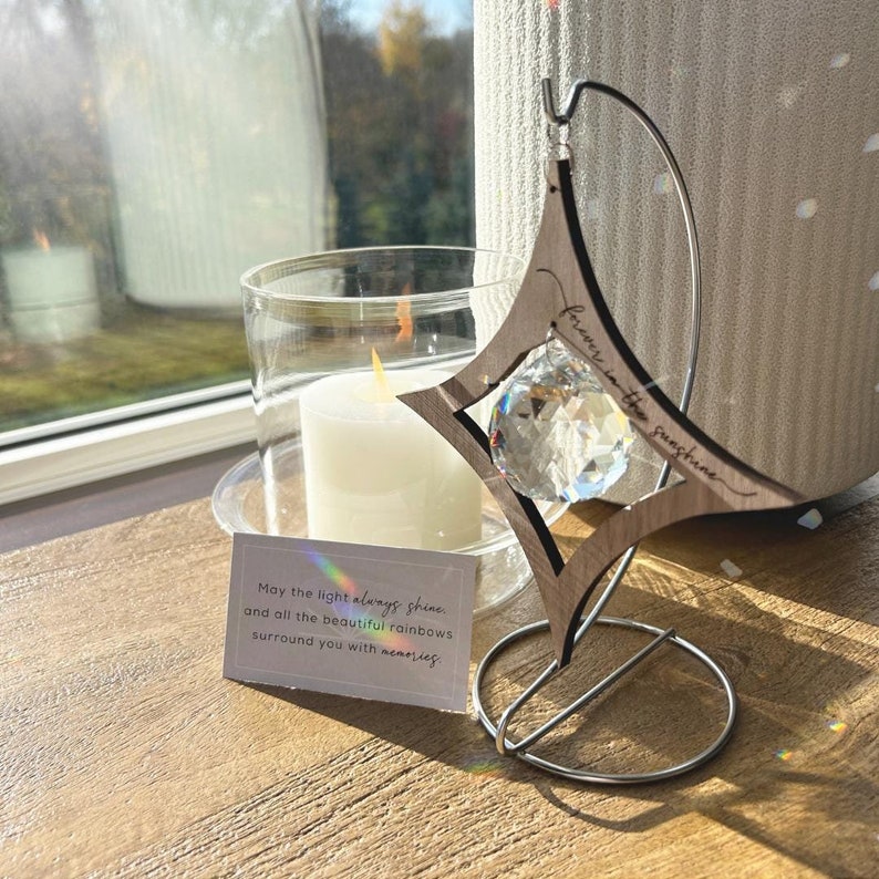 K&ouml;nnte beinhalten: Ein sternf&ouml;rmiger Kristall-Sonnenf&auml;nger aus Holz mit dem eingravierten Text "forever in the sunshine". Der Sonnenf&auml;nger h&auml;ngt an einem Metallst&auml;nder und hat einen Kristall-Prisma, der in der Mitte h&auml;ngt. Eine kleine Karte mit dem Text "May the light always shine and all the beautiful rainbows surround you with memories." liegt auf dem Tisch unter dem Sonnenf&auml;nger.