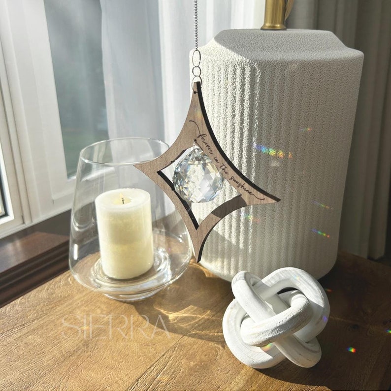 K&ouml;nnte beinhalten: Ein h&ouml;lzerner sternf&ouml;rmiger Kristall-Sonnenf&auml;nger mit dem Text "Forever in the sunshine", der an einer silbernen Kette h&auml;ngt. Der Sonnenf&auml;nger h&auml;ngt vor einer wei&szlig;en, strukturierten Vase und einem wei&szlig;en Keramik-Knoten.