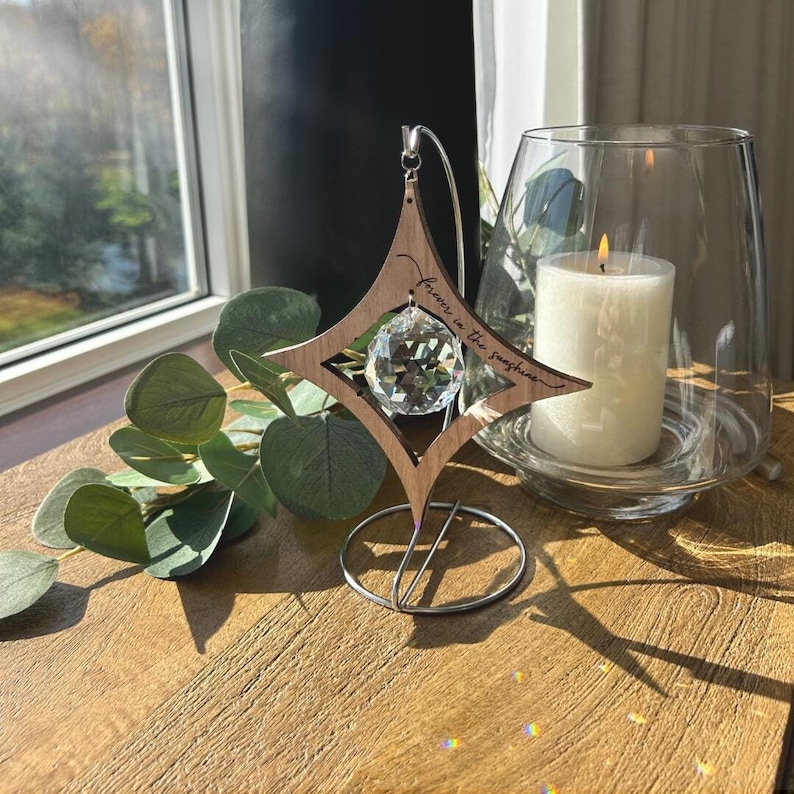 K&ouml;nnte beinhalten: Ein sternf&ouml;rmiger Kristall-Sonnenf&auml;nger aus Holz mit dem eingravierten Text "Forever in the sunshine". Der Sonnenf&auml;nger h&auml;ngt an einem Metallst&auml;nder und hat eine klare Kristallkugel in der Mitte.