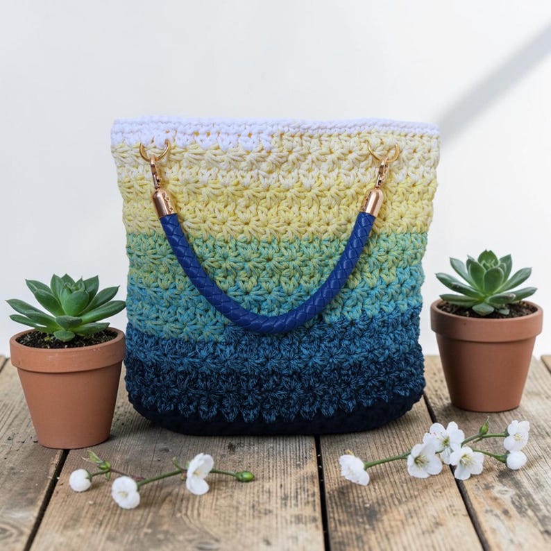 May include: A crocheted tote bag with a gradient of colors from white to yellow, green, and dark blue. The bag has a dark blue braided handle and gold-colored hardware. Two small potted succulents and white flowers are on either side of the bag.