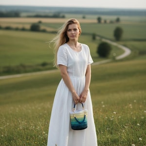 May include: A woman in a white dress holds a small, colorful crocheted handbag with blue, yellow, and green hues. The bag has a blue handle. The scene is set in a field with a winding road in the background.
