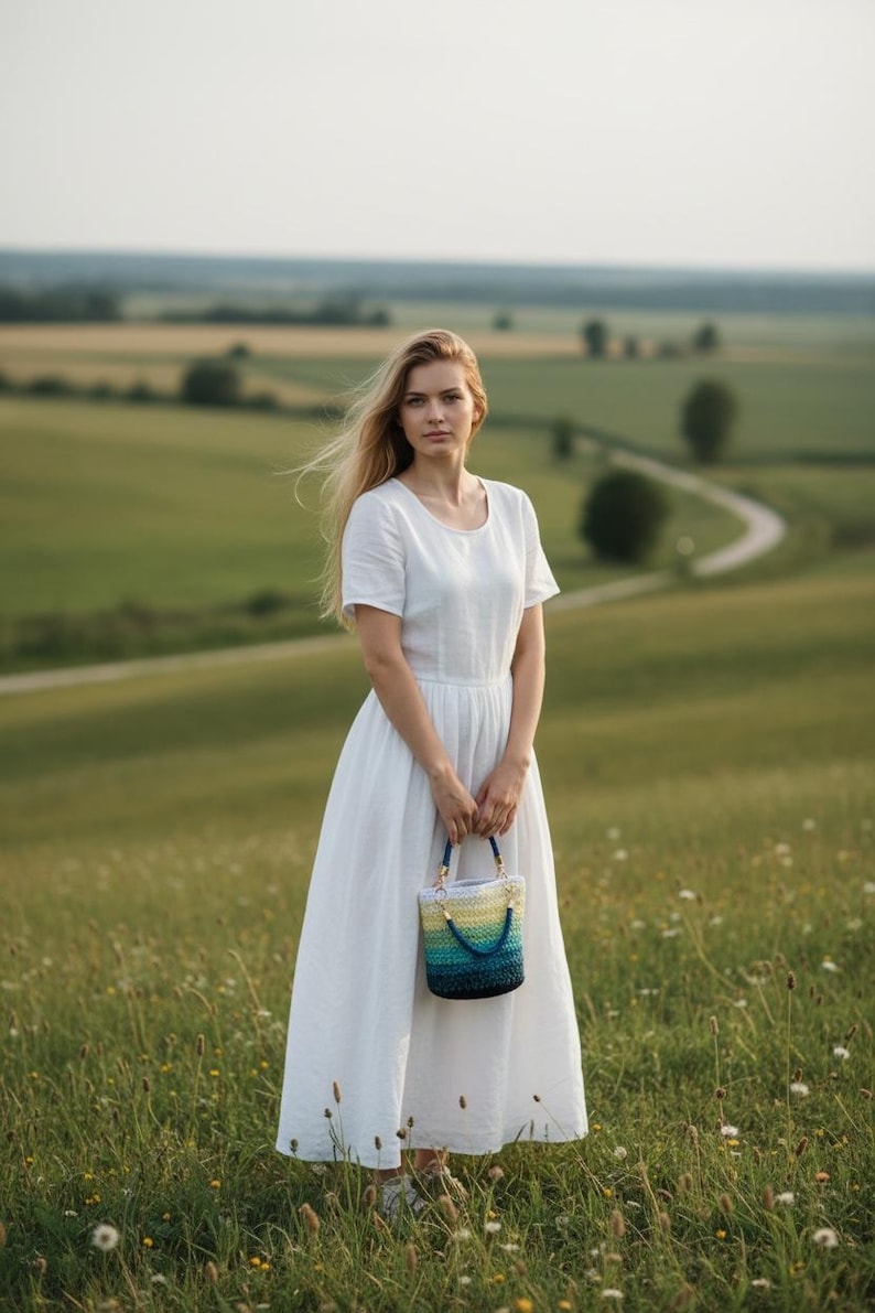 May include: A woman in a white dress holds a small, colorful crocheted handbag with blue, yellow, and green hues. The bag has a blue handle. The scene is set in a field with a winding road in the background.