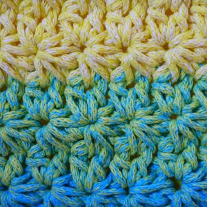 May include: Close-up of a crocheted fabric with a floral pattern. The color scheme transitions from yellow to turquoise, creating a gradient effect. The texture is dense and detailed, showcasing the intricate stitch work.