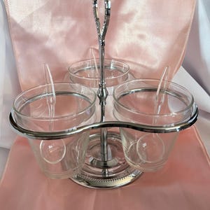 May include: A silver-toned metal condiment server featuring three clear glass bowls held in a tiered frame. The central vertical handle has a decorative loop at the top, and each glass bowl contains a small, clear plastic serving spoon.