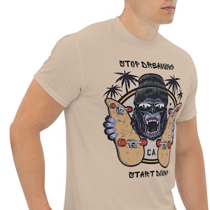 May include: A beige t-shirt featuring a graphic of a gorilla wearing sunglasses and a beanie, holding two skateboards. The text "STOP DREAMING START DOING" is printed on the shirt.