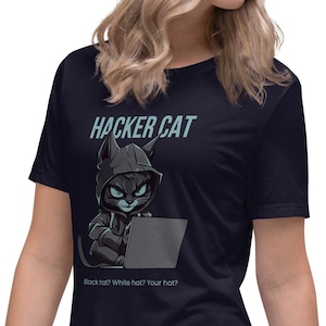 Op de afbeelding: Een donkerblauw t-shirt met een afbeelding van een kat met een hoodie die een laptop gebruikt. De tekst "Hacker Cat" staat boven de kat en de tekst "Black hat? White hat? Your hat?" staat onder de kat.