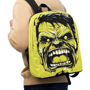Op de afbeelding: Een limoengroene rugzak met een afbeelding van een brullend, geelgroen Hulk-gezicht met zwart haar en witte tanden. De rugzak heeft zwarte schouderbanden en een zwart handvat aan de bovenkant. De persoon op de foto draagt een beige trui.