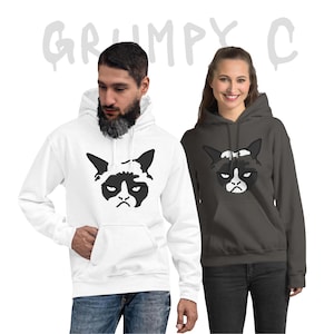 Peut inclure: Deux sweats à capuche, un blanc et un gris anthracite, chacun avec un dessin noir et blanc d'un visage de chat grincheux. Le mot "GRUMPY C" est écrit en gris au-dessus des sweats à capuche. Les deux sweats ont une poche avant.