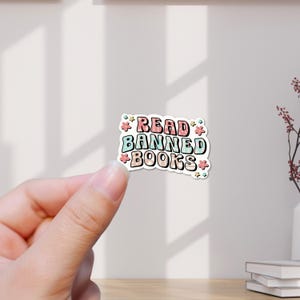 Könnte beinhalten: Ein Aufkleber mit dem Text "READ BANNED BOOKS" in einer Retro-Schriftart, umgeben von kleinen floralen Akzenten. Der Aufkleber hat einen weißen Hintergrund und wird von einer Hand gehalten.