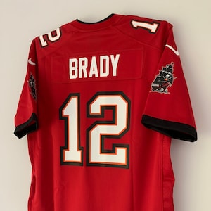 May include: Red football jersey with the name "BRADY" and the number "12" on the back. The jersey has a black and white pirate ship logo on the right sleeve.