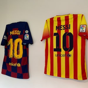 以下が含まれることがあります： 「メッシ」という名前と背番号「10」が書かれたサッカーのユニフォームが2着。左側のユニフォームは、青と赤の市松模様。右側のユニフォームは、黄色と赤の縦縞。両方のユニフォームには、下部に「ユニセフ」の文字が印刷されています。