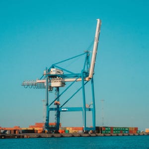 May include: A tall blue and white harbour crane set against a clear blue sky. The crane is positioned over water, with colourful shipping containers visible on a dock in the distance. The scene is bright and sunny.