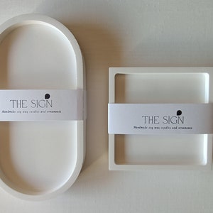 Peut inclure: Deux plateaux en céramique blanche, l'un ovale et l'autre carré, avec le texte "THE SIGN Handmade soy wax candles and ornaments" sur chacun.