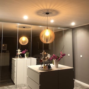 Può includere: Un mobile da bagno bianco con due cassetti e un piano in bianco. Il mobile da bagno è di fronte a una parete a specchio con due luci a sospensione a forma di globo in legno appese al soffitto. Le luci sono realizzate con doghe di legno naturale e hanno una luce calda e invitante. Sul piano ci sono oggetti decorativi, tra cui una piccola statuina bianca, un vaso con fiori rosa e un vassoio con varie bottiglie e barattoli.