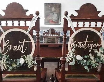 Op de afbeelding: Twee houten stoelen met witte linten en groen. Elke stoel heeft een rond houten bord met de woorden "Braut" en "Brautigan" in wit.