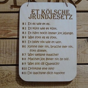 Peut inclure: Panneau en bois avec le texte "ET KÖLSCHE JRUNDJESETZ" et onze affirmations numérotées en allemand. Le panneau est rectangulaire avec des coins arrondis et un trou pour l'accrocher. Le texte est de couleur marron foncé.