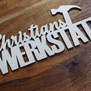 Può includere: Un cartello in legno bianco con il testo "Christians Werkstatt" e un martello stilizzato sopra il testo.