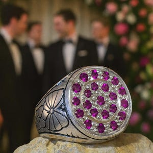 Puede incluir: Un anillo plateado con un diseño floral en el lateral y un grupo de pequeñas gemas redondas de color magenta en el centro. El anillo se muestra sobre una roca, con el texto "Jerusalem Rings" en la parte superior.