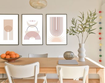 May include: Three framed abstract art prints with geometric shapes in shades of pink, red, and brown. The prints are hung on a white wall above a wooden dining table with two white chairs.