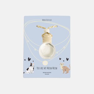 Op de afbeelding: Een witte glazen diffuser met een houten dop en een touwtje om op te hangen. De diffuser bevindt zich op een blauwe kaart met de tekst "You are my meow meow" en twee cartoonkatten. De kaart vermeldt ook "Aroma Moods Scented" en "Mood Diffuser".